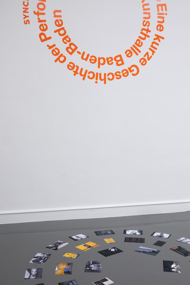 Eine Ausstellungsansicht von SYNCH03: An Imaginary Audience. Das Bild zeigt den Titel der Ausstellung in einer orangefarbenen Textspirale an der Wand angebracht und unmittelbar davor - wie gespiegelt - eine gleich große Spirale aufgedruckt auf dem Boden. Diese Spirale besteht aus kleinen rechteckigen Fotografien, welche die Werke der Ausstellung zeigen.