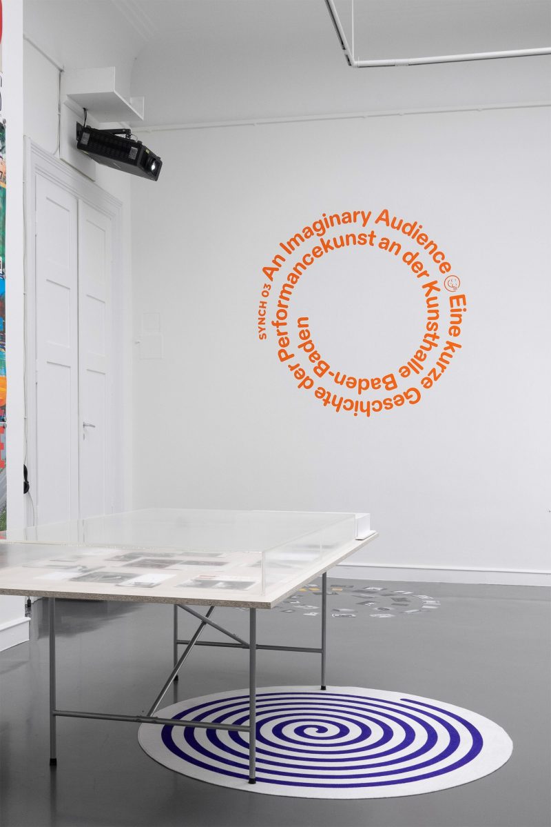 Eine Ausstellungsansicht von SYNCH03: An Imaginary Audience. Das Bild zeigt den Blick in den Ausstellungsraum, wo im Hintergrund der Titel der Ausstellung in einer orangefarbenen Textspirale an der Wand angebracht ist. Im Vordergrund liegt ein runder, weißer Teppich auf dem Boden, auf dem eine lilafarbene Spirale gedruckt ist. Halb darauf steht ein Tisch mit dünnen Stahlbeinen und einer Haube aus Plexiglas, darunter sind Archivalien zu erkennen, wie Poster, DVDs, Einladungskarten und Fotos.